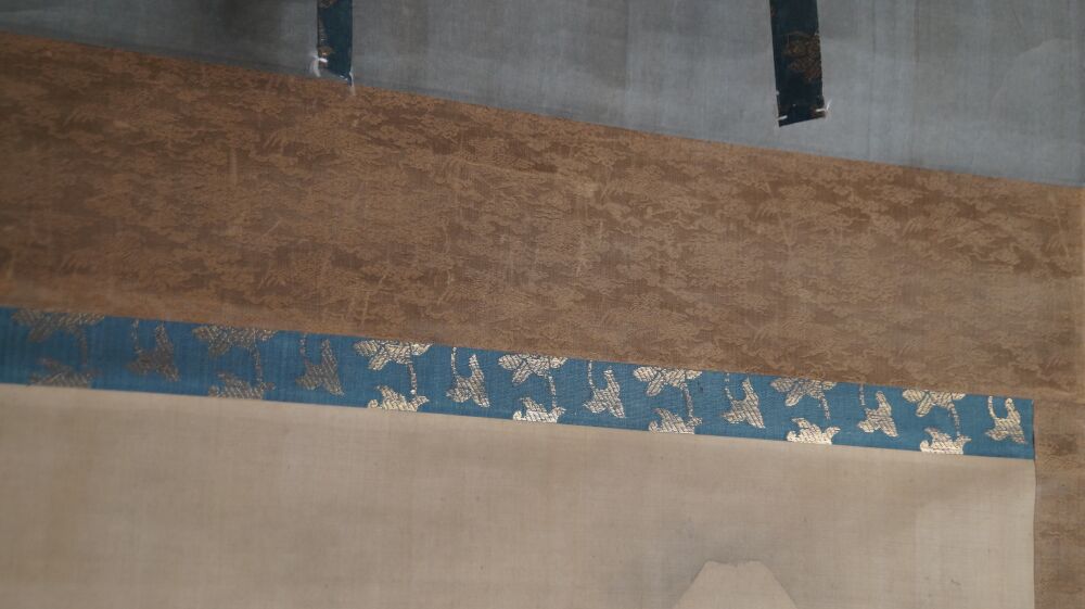 Vista dettagliata del montaggio in seta con motivo di pini marroni e nastro broccato con gru blu del kakemono Fuji tra le vette avvolte dalle nuvole