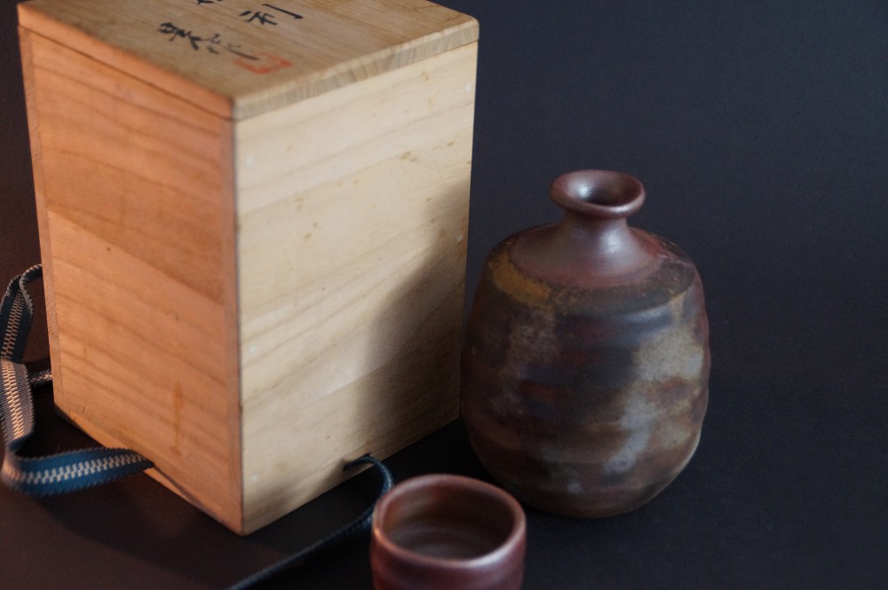 Die Sake Flasche mit Guinomi neben der signierten Aufbewahrungsbox mit Kordel