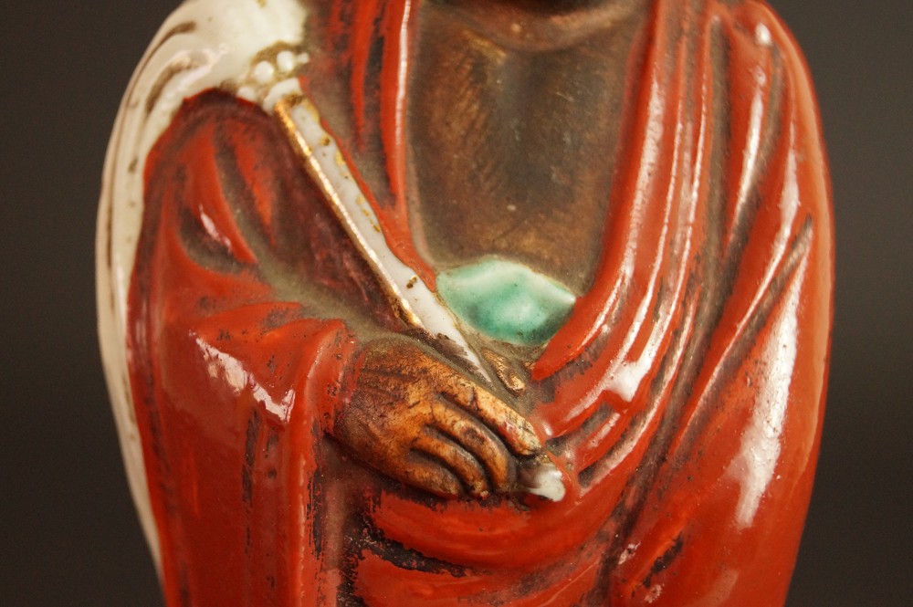 Bodhidharma Daruma Figur aus rotem Kutani-Porzellan Nahaufnahme der gefalteten Haende mit Gewandfalten Motiv Nahaufnahme