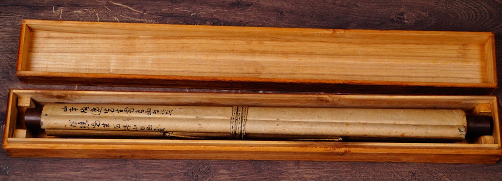 Zusammengerolltes und mit Kordel verschnuertes Kakemono Reiher auf Fels mit Chrysanthemen von Yamamoto Baiitsu mit sichtbarer Tuschebeschriftung auf dem Einschlagpapier in der geoeffneten Aufbewahrungsbox