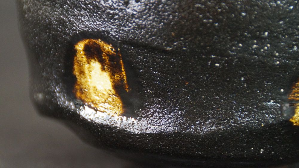 Primer plano extremo de la mancha de esmalte dorado sobre el esmalte negro del chawan de cerámica Raku