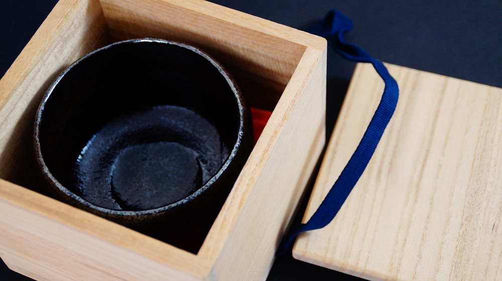 Chawan aus Raku-Keramik von Shoraku Sasaki liegend in der geoeffneten Aufbewahrungsbox mit dunkelblauer Verschnuerung