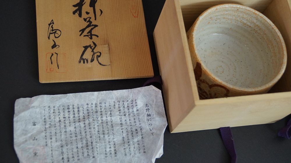 Chawan aus Shino-Keramik in der geoeffneten Aufbewahrungsbox neben dem Kastendeckel und dem Echtheitszertifikat zum Toyama Ofen