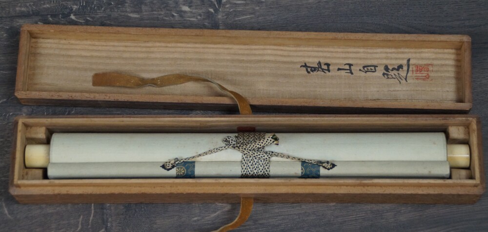 Zusammengerolltes Kakemono Begegnung am Rand des Wasserfalls mit elfenbeinfarbenen Rollenenden in der geoeffneten Aufbewahrungsbox mit Tuschebeschriftung des Deckels
