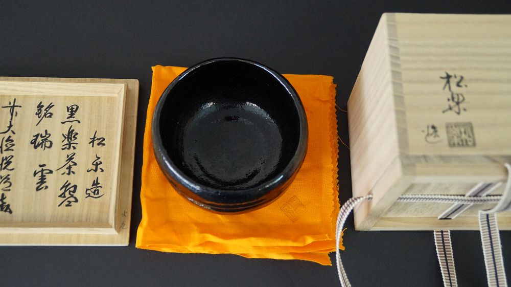 Czarka do herbaty chawan na pomarańczowej tkaninie obok opisanej pokrywy pudełka Kuroraku Chawan mei Zuiun oraz pudełka do przechowywania