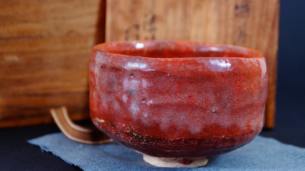 Vista lateral del cuenco de té de cerámica Raku de Sairi Hotoku junto a la caja de conservación sobre tela azul