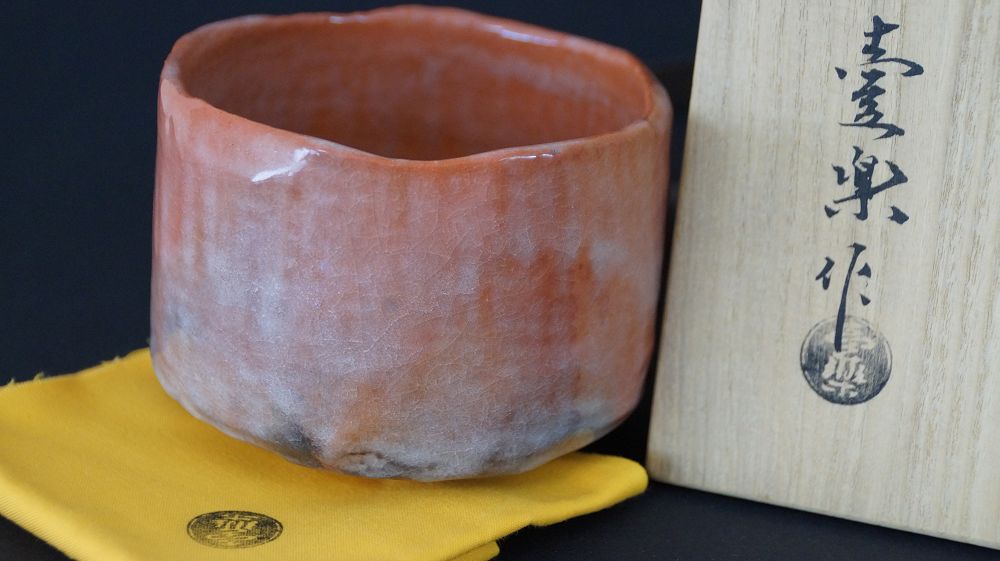 Weitere Ansicht der Chawan Teeschale auf gelbem Tuch neben der Aufbewahrungsbox mit der Aufschrift Raku
