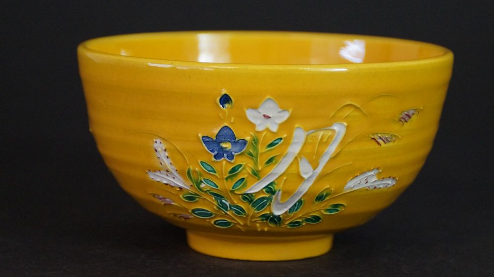Weitere Ansicht der Chawan aus Kyoto-Keramik mit weiss aufgetragenem Schriftzeichen zwischen bemalten Blueten