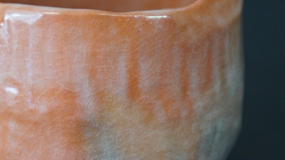 Nahaufnahme des unregelmaessig geformten Randes mit glaenzender orangefarbener Glasur der Chawan Teeschale aus Raku-Keramik